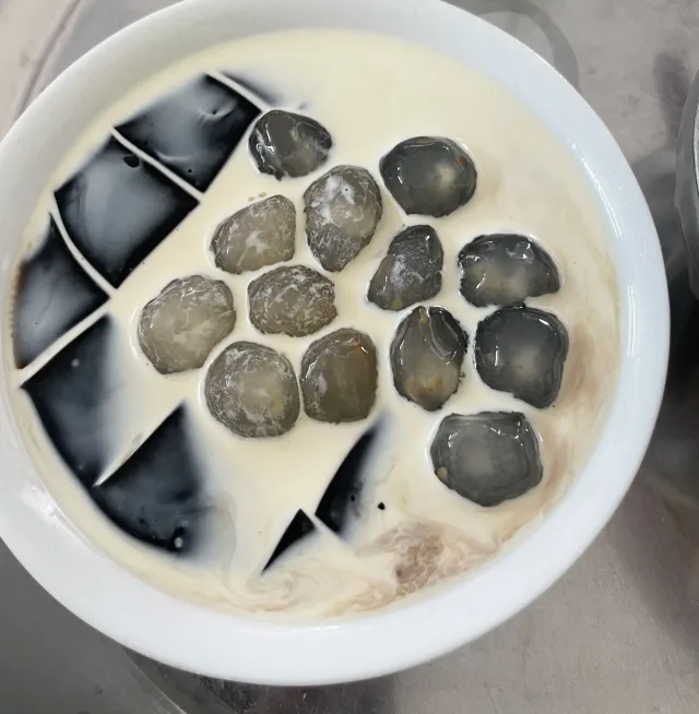 A bowl of chè with black grass-jelly cubes and round pieces in a pale liquid