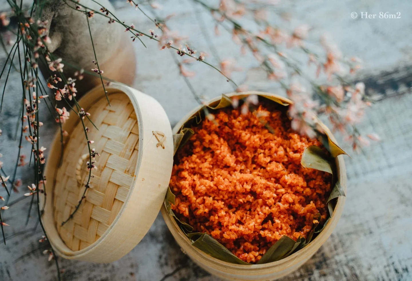 Red gấc sticky rice in a bamboo steamer with a floral branch
